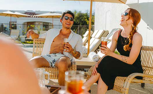 Man and woman chat and hold drinks while sitting on a chairs by the Miralina pool under an umbrella